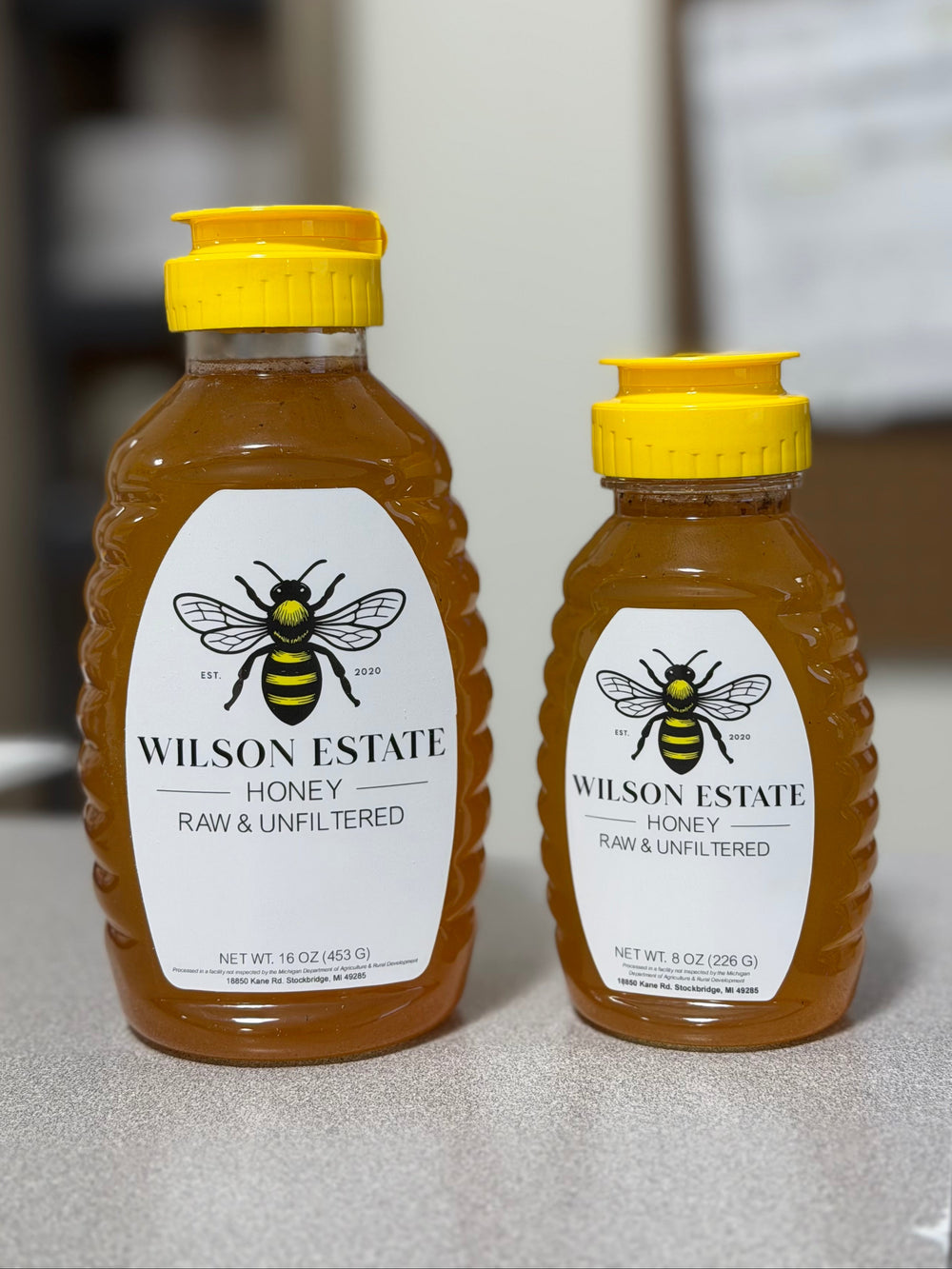 Two bottles of Wilson Estate Raw & Unfiltered Honey, one 16 oz and one 4 oz, standing on a gray countertop with a blurred indoor background.